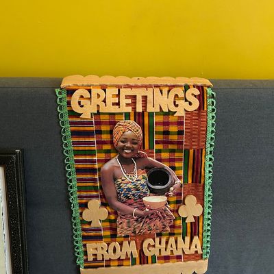 Sale Photo Thumbnail #121: Vintage "Greetings from Ghana" souvenir wall hanging featuring a colorful Kente cloth background, wooden lettering, and a central image of a woman in traditional attire.