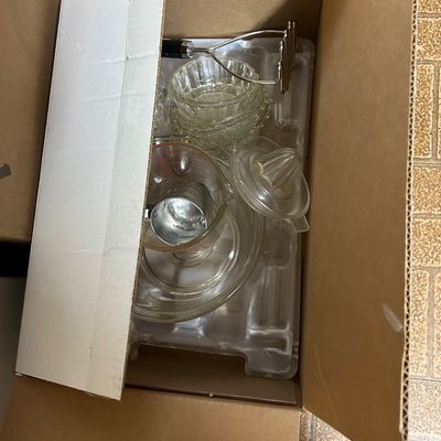 Sale Photo Thumbnail #83: Cardboard box containing assorted vintage kitchenware, including a glass measuring cup, glass juicer, small glass bowls, and a metal potato masher.