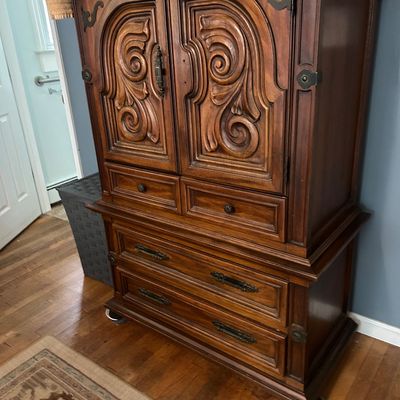 Sale Photo Thumbnail #105: Vintage Spanish Revival style wooden armoire featuring ornate carved cabinet doors, two small drawers, and two large lower drawers with decorative metal hardware.