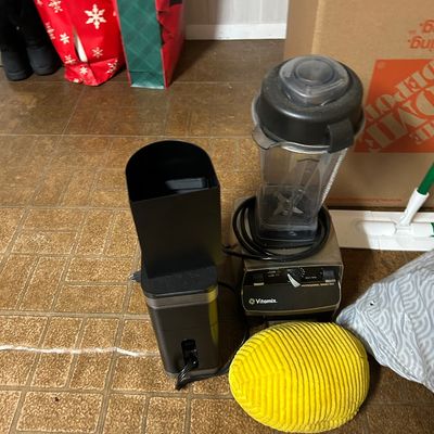 Sale Photo Thumbnail #85: Vitamix Professional Series 500 blender with a clear pitcher, shown alongside a black coffee grinder and a yellow corduroy cushion on a tiled floor.