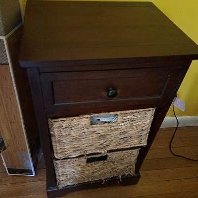 Sale Photo Thumbnail #124: Dark wood nightstand featuring a single drawer with a black knob and two open shelves holding woven wicker storage baskets.