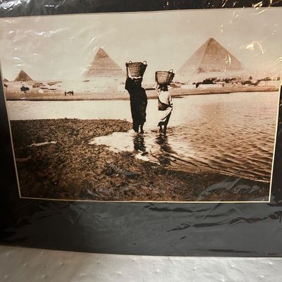 Sale Photo Thumbnail #57: Matted vintage sepia-toned photograph depicting two figures carrying baskets on their heads near water, with the Pyramids of Giza in the background.