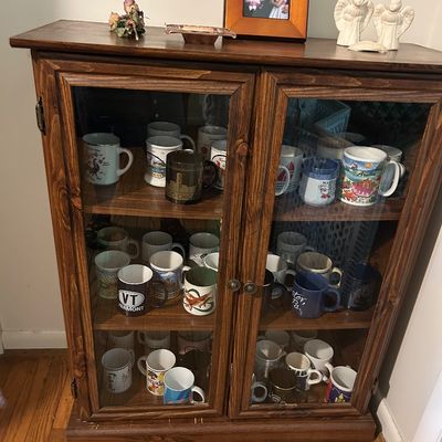 Sale Photo Thumbnail #149: Rustic pine display cabinet with double glass doors, housing a collection of souvenir and novelty coffee mugs.