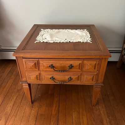 Sale Photo Thumbnail #145: Mid-century modern wooden end table featuring two drawers with ornate metal pulls and geometric carved detailing, topped with a white lace doily.