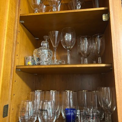 Sale Photo Thumbnail #17: Assorted glassware collection including martini glasses, wine goblets, highball glasses with gold rims, and novelty shot glasses, displayed inside a wooden cabinet.