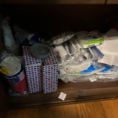 Sale Photo Thumbnail #152: Assortment of disposable party supplies, including paper plates, plastic cups, napkins, and plastic cutlery, stored inside a wooden cabinet.