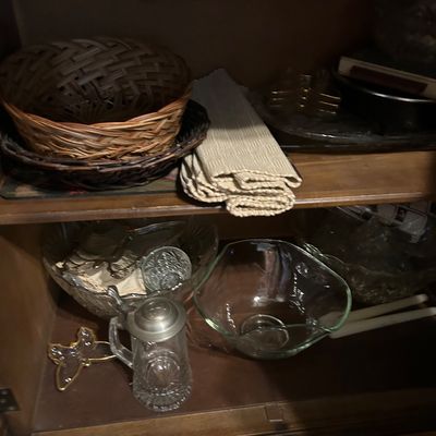 Sale Photo Thumbnail #151: Assorted glassware and home decor items, including woven baskets, a glass stein with a metal lid, and various clear glass serving bowls, displayed inside a wooden cabinet.