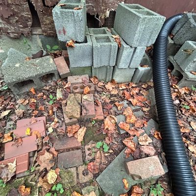 Sale Photo Thumbnail #3: Assorted concrete cinder blocks, red bricks, and a length of black corrugated drainage pipe scattered outdoors.