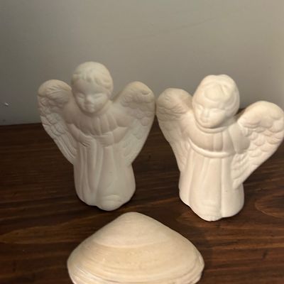 Sale Photo Thumbnail #148: Pair of unpainted ceramic bisque angel figurines alongside a large natural clam shell on a wooden surface.