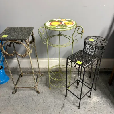 Sale Photo Thumbnail #182: Assorted metal plant stands in various styles and heights, including one with a granite top, a green stand with a hand-painted fruit tile top, and two black metal stands.
