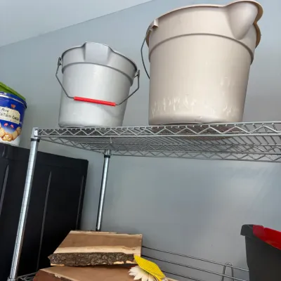 Sale Photo Thumbnail #153: Two plastic utility buckets, one silver with a red handle grip and one beige, displayed on a wire shelving unit above two live-edge wood slabs.