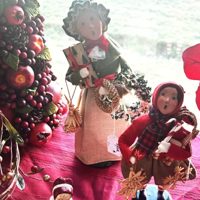 Sale Photo Thumbnail #56: Vintage Byers' Choice Carolers figurines, including a woman in a green coat holding a gift and a child in a red coat holding a candy cane, displayed alongside festive holiday decor.