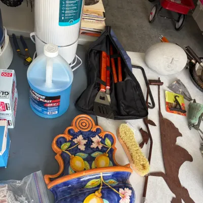 Sale Photo Thumbnail #147: An assortment of household items including a hand-painted ceramic lemon wall pocket, a Homedics ultrasonic humidifier, a set of BBQ tools in a zippered case, and a decorative stone face.