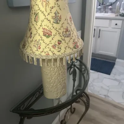 Sale Photo Thumbnail #119: Vintage-style table lamp featuring a textured cream ceramic base and a yellow fabric shade with a chinoiserie pattern and tassel trim, displayed on a wrought iron console table.