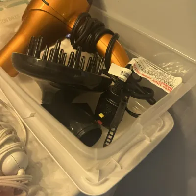 Sale Photo Thumbnail #142: Metallic orange hair dryer with a black diffuser attachment and power cord, stored inside a clear plastic bin.