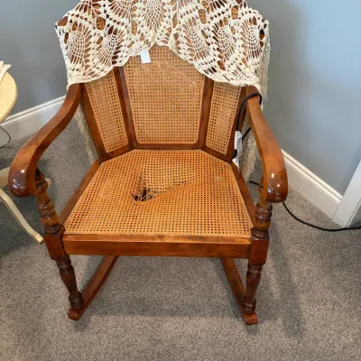 Sale Photo Thumbnail #8: Vintage wooden rocking chair featuring a cane back and seat, with a notable tear in the seat cane, draped with a white crocheted lace shawl.