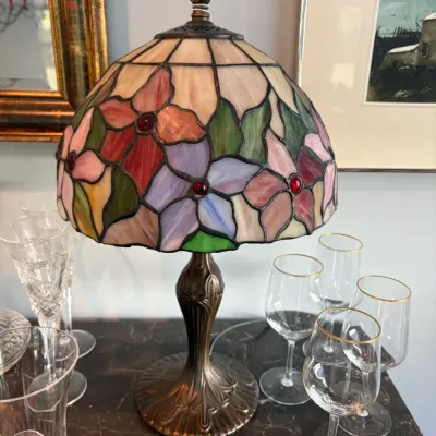 Sale Photo Thumbnail #120: Tiffany-style table lamp featuring a stained glass shade with a colorful floral motif and red glass gems, set on an ornate bronze-finished metal base.
