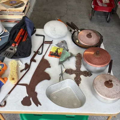 Sale Photo Thumbnail #148: Assortment of vintage kitchenware and garden decor, featuring aluminum pots with copper-colored lids, a cast iron cross, a carved stone face, and a rustic metal fairy silhouette.