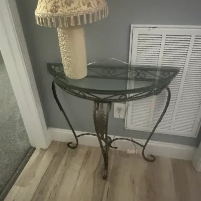 Sale Photo Thumbnail #83: Demilune console table featuring a wrought iron frame with scrollwork details and a glass top, paired with a ceramic table lamp with a fringed fabric shade.