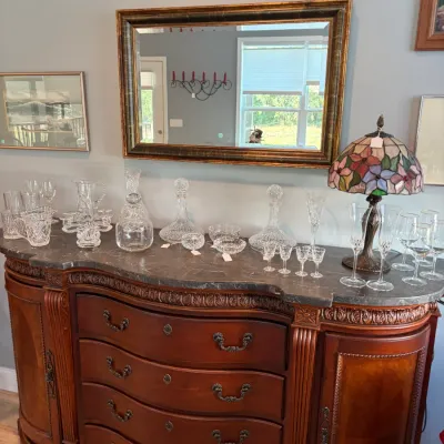 Sale Photo Thumbnail #84: Ornate traditional wooden sideboard with a dark marble top, featuring curved drawers, side cabinets, and detailed carvings, styled with various crystal glassware and a Tiffany-style stained glass lamp.