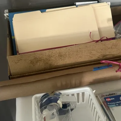 Sale Photo Thumbnail #164: Assorted office supplies including manila file folders in a cardboard box, a roll of brown wrapping paper, and tape dispensers in a small white plastic basket.