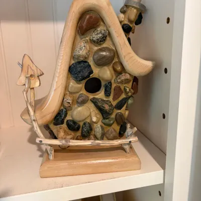 Sale Photo Thumbnail #183: Whimsical rustic wooden birdhouse sculpture inlaid with various river stones, featuring a curved roofline and miniature twig fence detail.