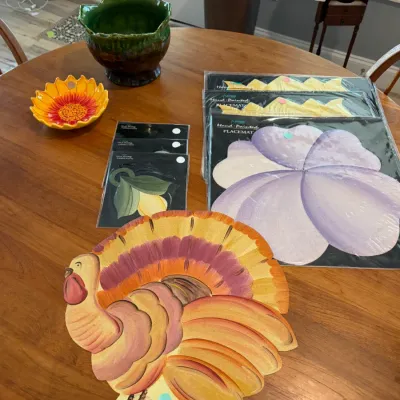 Sale Photo Thumbnail #63: Assortment of hand-painted tabletop decor, featuring a wooden turkey cutout, packaged floral placemats and napkin rings, a sunflower dish, and a green glazed ceramic planter.