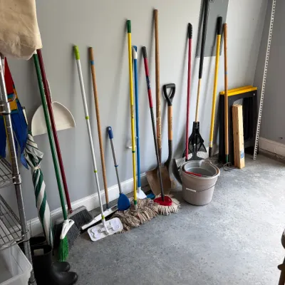 Sale Photo Thumbnail #168: Assortment of household cleaning and yard tools, including brooms, mops, shovels, a rake, and a plastic bucket, leaning against a garage wall.