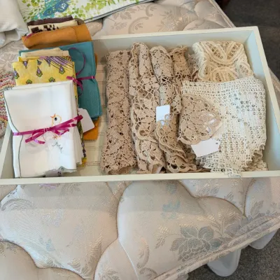 Sale Photo Thumbnail #124: Assorted vintage linens arranged in a white tray, including embroidered napkins tied with pink ribbon, colorful folded fabrics, and various crocheted lace doilies and runners.