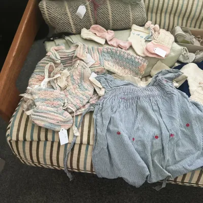 Sale Photo Thumbnail #144: Assortment of vintage baby clothing, including a blue and white striped dress, hand-knit pastel sweaters, booties, and socks, displayed on a striped sofa.