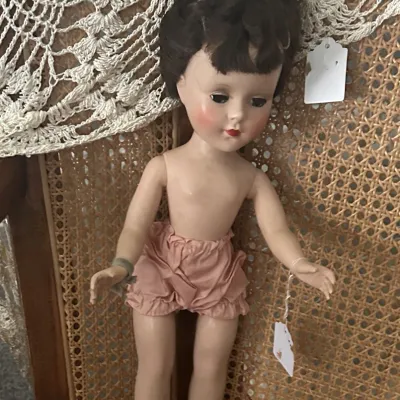 Sale Photo Thumbnail #5: Vintage 1950s hard plastic walker doll with dark brown hair, sleep eyes, and pink bloomers, displayed on a cane-back chair.