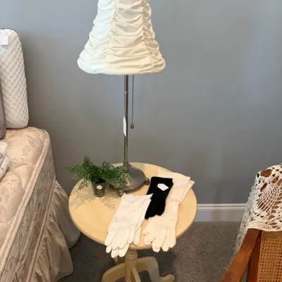 Sale Photo Thumbnail #116: Silver-toned table lamp with a ruched white fabric shade, displayed on a cream-colored round wooden side table alongside pairs of vintage dress gloves.