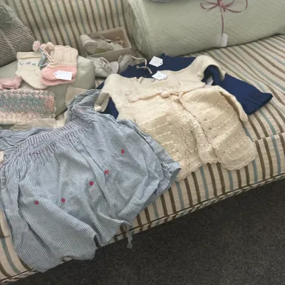 Sale Photo Thumbnail #145: Assortment of vintage baby clothing and blankets, including hand-knit sweaters, booties, and a striped romper, displayed on a striped sofa.