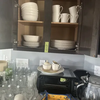 Sale Photo Thumbnail #101: Collection of white ceramic dinnerware, including bowls, plates, and mugs, displayed in a dark wood cabinet alongside various glassware and kitchen appliances.