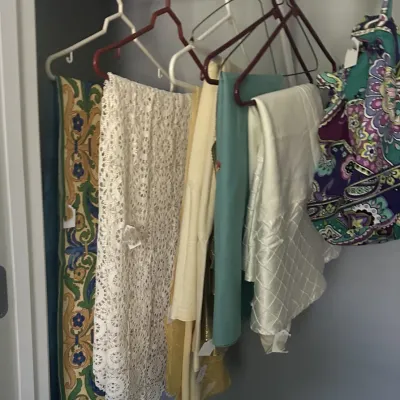 Sale Photo Thumbnail #125: Assorted vintage linens, including a white lace table runner, a patterned floral runner, and various solid-colored tablecloths, hanging on hangers in a closet.