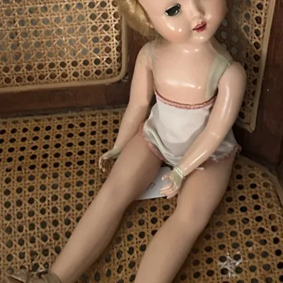Sale Photo Thumbnail #6: Vintage mid-century hard plastic doll with blonde hair, sleep eyes, and original shoes, seated on a cane-backed chair.