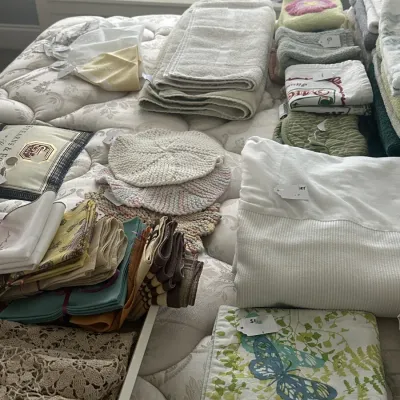 Sale Photo Thumbnail #129: Assortment of household linens, including folded towels, cloth napkins, crocheted items, and a butterfly-patterned placemat, arranged on a mattress.