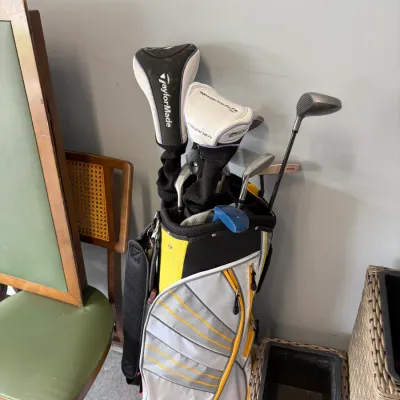 Sale Photo Thumbnail #181: TaylorMade golf bag in white, grey, and yellow, containing a set of golf clubs including TaylorMade woods with headcovers and various irons.