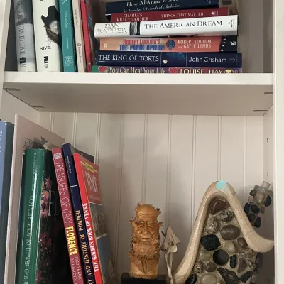 Sale Photo Thumbnail #39: A collection of books, a small bust of George Bernard Shaw, and a decorative stone-accented birdhouse on white wooden shelves.