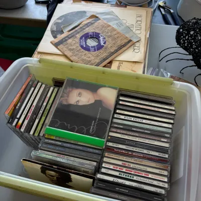 Sale Photo Thumbnail #59: Plastic storage bin filled with assorted music CDs from the 1990s and 2000s, alongside a stack of vintage vinyl records.