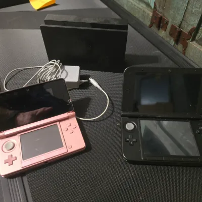 Sale Photo Thumbnail #184: A collection of Nintendo gaming hardware. Includes one metallic pink Nintendo 3DS console, one black Nintendo 3DS XL console, a Nintendo 3DS AC adapter charger, and a black Nintendo Switch docking station. Items show some light surface wear and dust from 