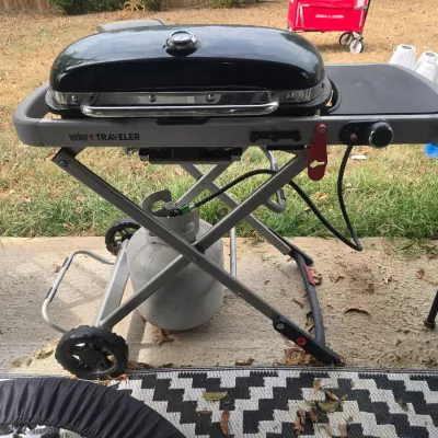 Sale Photo Thumbnail #212: A Weber Traveler portable gas grill in black and silver. Features a folding scissor cart design with wheels for easy transport, a built-in lid thermometer, and a side table. Includes a connected propane tank. Appears to be in gently used condition with mi