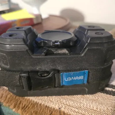Sale Photo Thumbnail #187: A rugged, heavy-duty Braven portable Bluetooth speaker in a black shockproof casing. Features a built-in mounting strap with a plastic buckle and a blue Braven logo tag. The item shows some light dust and surface wear from storage but appears intact. Func