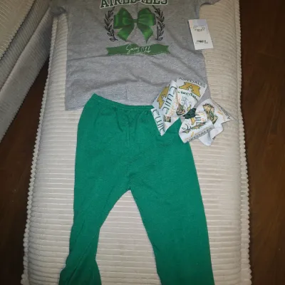 Sale Photo Thumbnail #195: A children's clothing set featuring the Alma Airedales. Includes a grey short-sleeve graphic t-shirt with a green bow design, green ruffle-hem pants, and a matching patterned hair bow. The shirt is new with tags. 5t