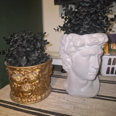 Sale Photo Thumbnail #199: A pair of decorative planters featuring classical motifs. Includes one white ceramic or plaster-style male bust head planter and one ornate gold-toned pot adorned with cherub faces. Both planters come styled with dark, faux eucalyptus-style foliage. In go