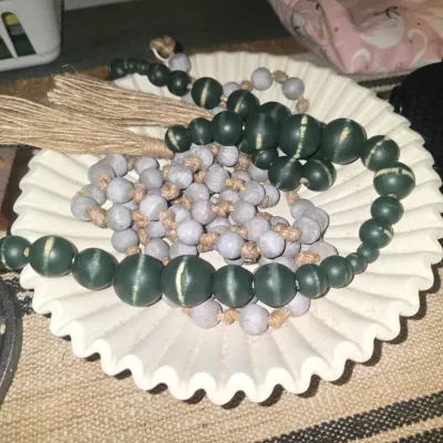 Sale Photo Thumbnail #200: A decorative off-white ceramic or plaster bowl featuring a fluted, wavy edge design. Inside the bowl are two decorative wooden bead garlands: one with larger dark green painted beads and jute tassels, and another with smaller light grey painted beads knot