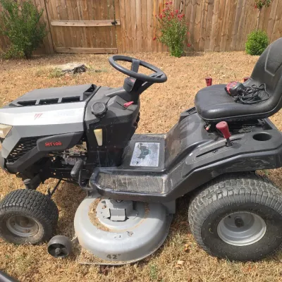 Sale Photo Thumbnail #204: Troy-Bilt MT100 riding lawn mower featuring a 42-inch cutting deck. Shows typical signs of outdoor use with some dust and grass clippings on the deck. Includes a battery charger resting on the seat. Tires appear in good condition with visible tread. No ho