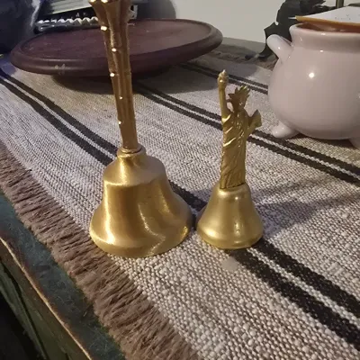Sale Photo Thumbnail #190: A pair of decorative gold-tone metal hand bells. One features a tall, textured handle, and the smaller bell features a Statue of Liberty handle. Both show some light surface wear and aging, but are in good overall condition.