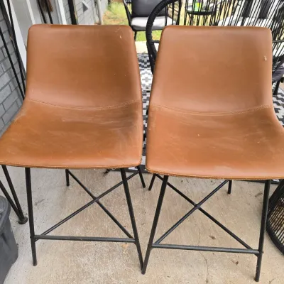Sale Photo Thumbnail #207: A pair of contemporary barstools featuring camel-colored faux leather upholstered seats with a curved silhouette. Supported by black metal legs with cross-brace detailing. Shows some light surface wear, dust, and minor scuffs from outdoor/patio use, but r