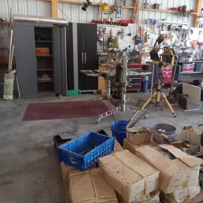 Workshop interior featuring various tools, storage cabinets, a radial arm saw on a mobile stand, and a pallet loaded with boxed inventory.
